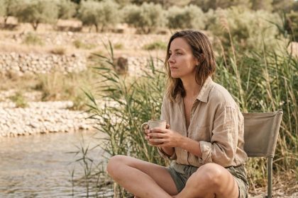 Femme en camping au bord d'une rivière en Provence, assise dans un fauteuil de camping avec une tasse, entourée de végétation méditerranéenne